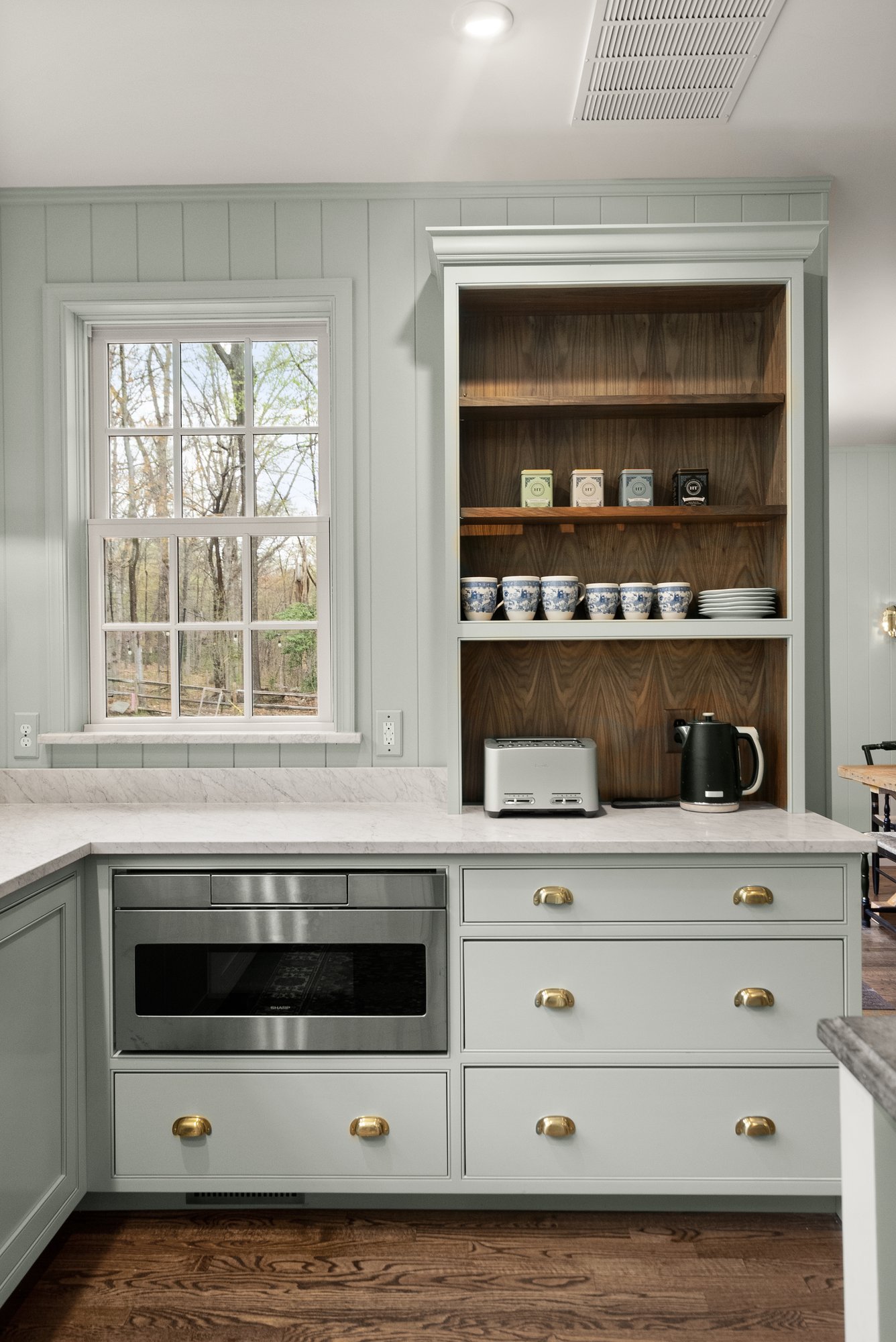 Open shelves in kitchen displaying coffee mugs, a toaster, a kettle, and wooded view through bright windows