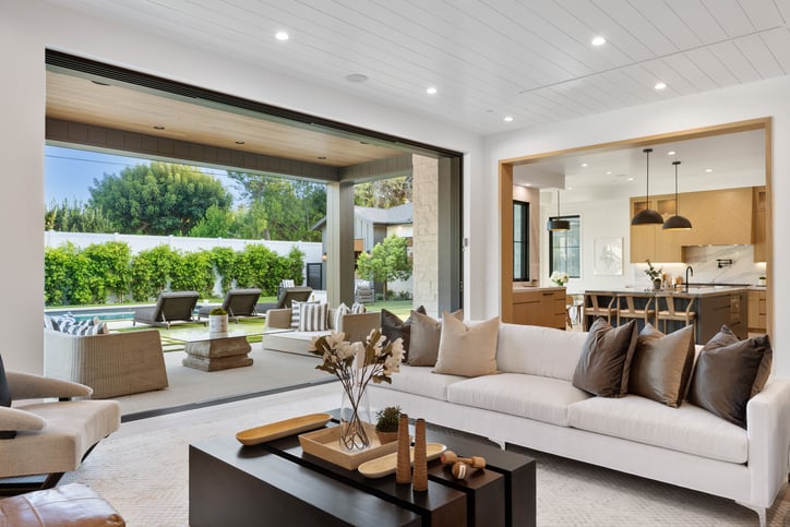 Modern living room with open sliding doors leading to a patio and garden area with lounge chairs and a pool.