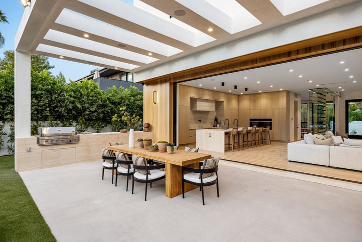 Modern open-plan kitchen and outdoor dining space with wooden furniture and greenery, featuring a seamless indoor-outdoor flow.