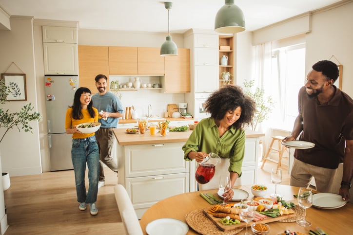 Group of people, diverse male and female friends preparing a meal and drinking cocktails together in domestic kitchen.